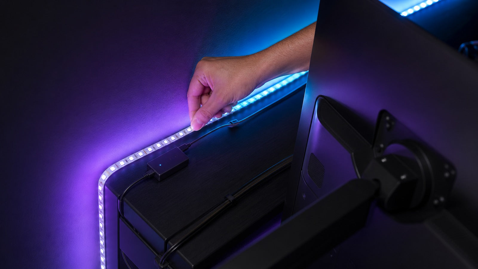 Hand installing an LED strip behind a monitor with purple and cyan bias lighting